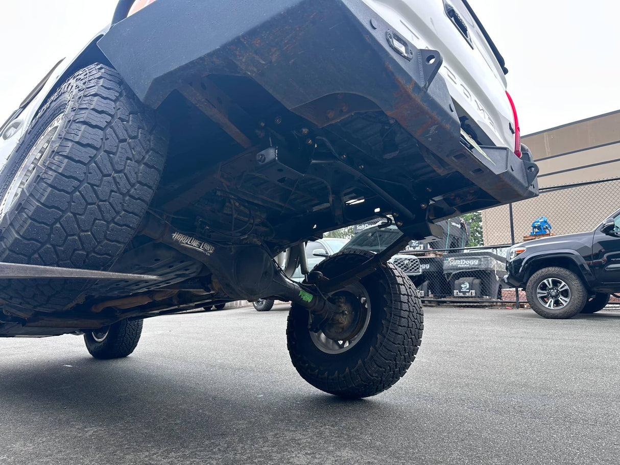 Close-up low-angle view of the undercarriage and rear suspension of a lifted black pickup truck, showing the rear tire, exhaust area, and mechanical components in an outdoor lot.