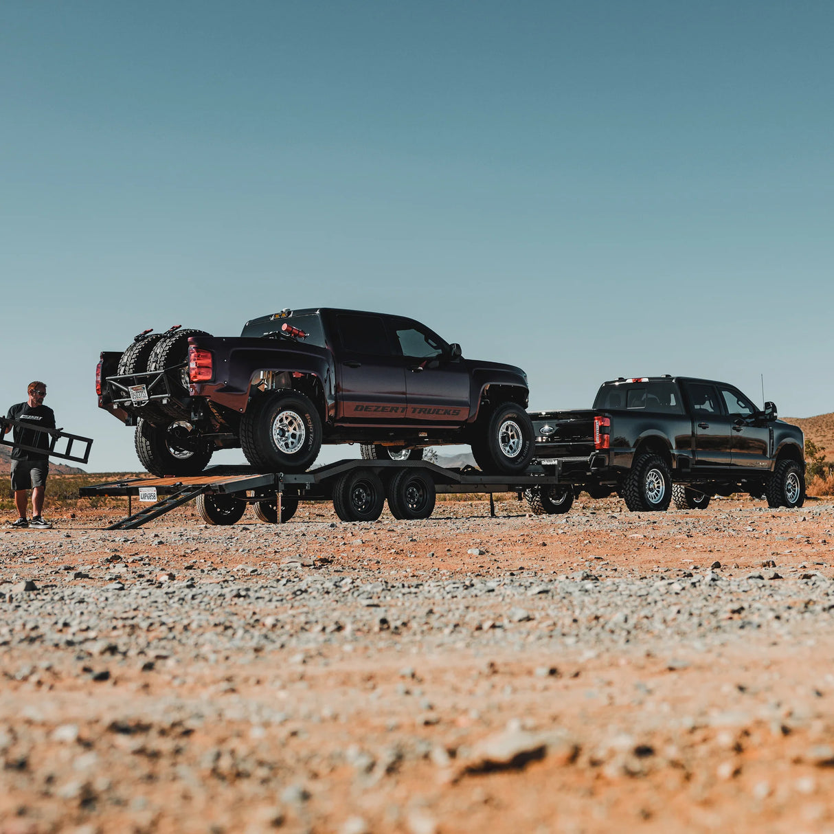Two pickup trucks on a trailer in a desert setting with a person standing nearby.