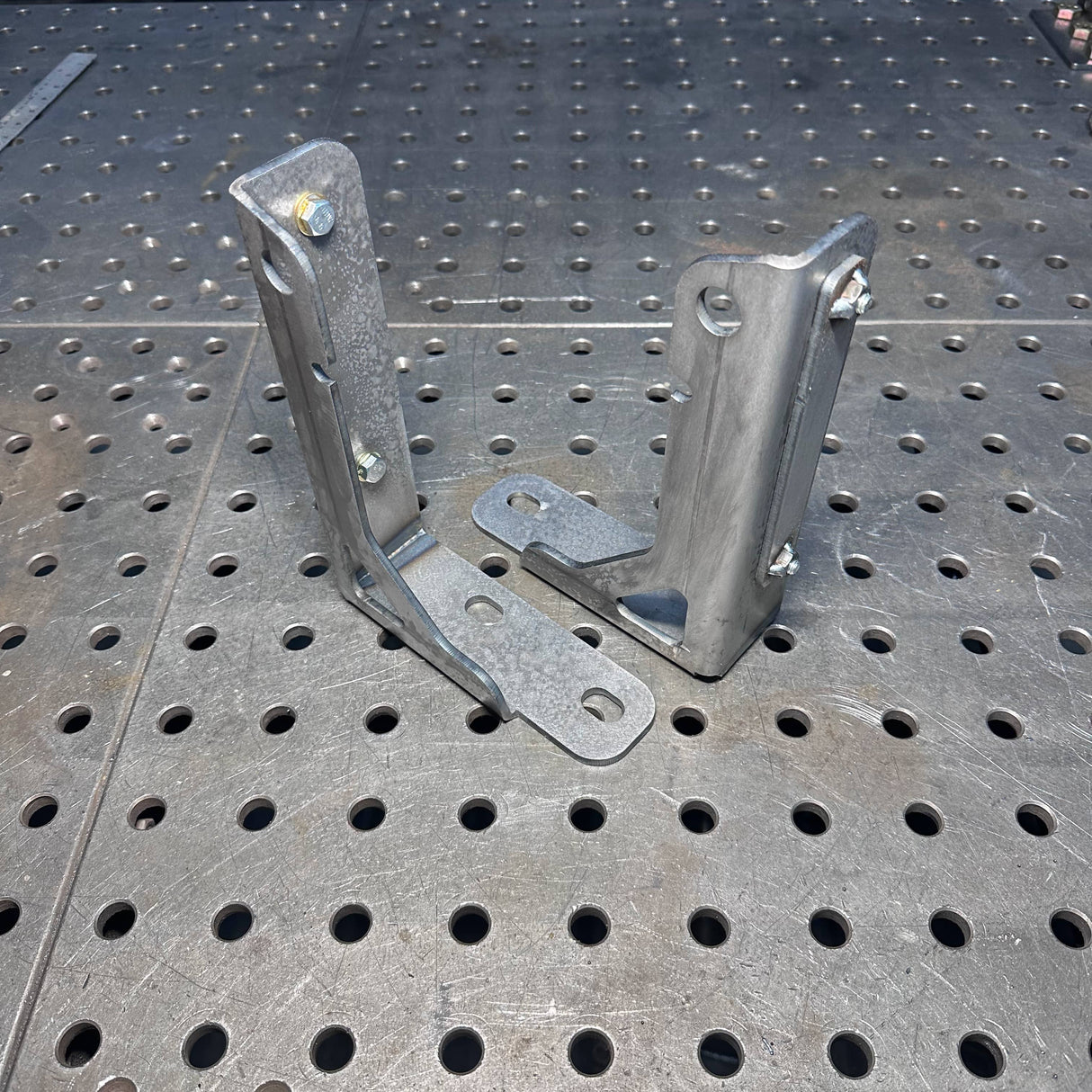 Two galvanized steel angle brackets with mounting plates, standing upright on a perforated metal workbench surface.