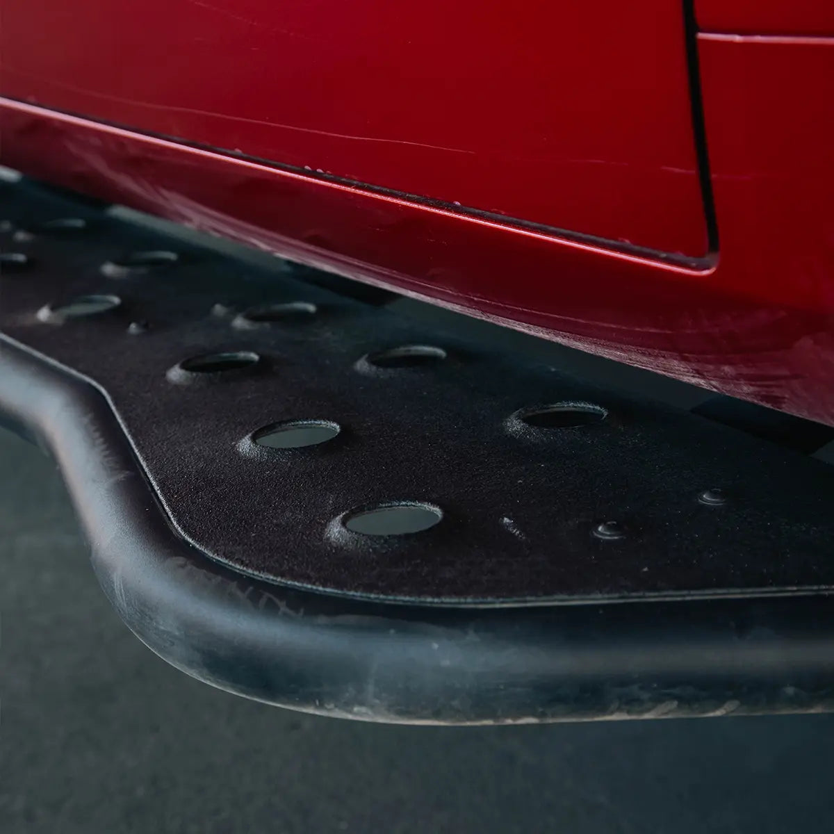 Close-up of a black metal vehicle side step running board mounted beneath a red car body panel.