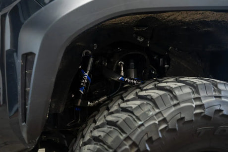 Close-up of a vehicle's suspension system showing black PEAK brand shock absorbers mounted inside the wheel well next to a large off-road tire.