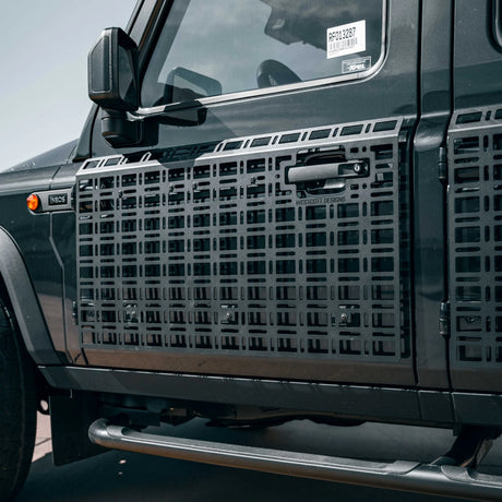 Black metal side step grates installed on the side of a dark gray off-road vehicle, featuring a grid pattern and a central handle cutout.