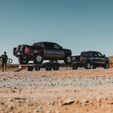 Two pickup trucks on a trailer in a desert setting with a person standing nearby.