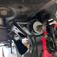Close-up of a black metal vehicle suspension control arm bolt and bushing assembly, showing a white gasket and a rusty bolt head, photographed in a workshop setting.