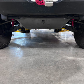 A close-up, low-angle view of a vehicle's undercarriage, showing a brushed metal skid plate on a concrete floor inside a garage.