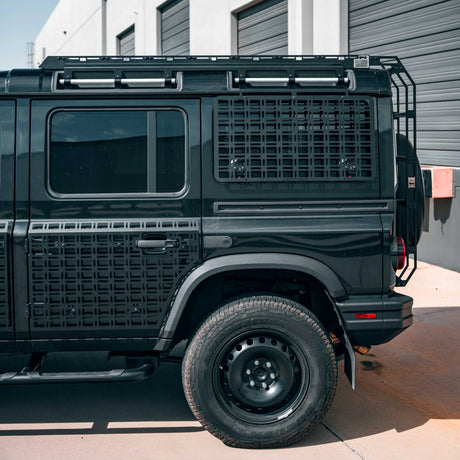 A black off-road vehicle with custom black metal grille side panels and a roof rack, parked outdoors in front of a gray building with garage doors.